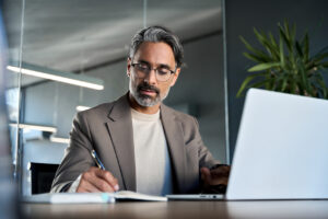 Senior marketing strategist writing notes while reviewing data on laptop, balancing human expertise with AI-enabled technology