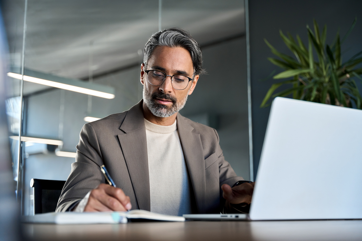 Senior marketing strategist writing notes while reviewing data on laptop, balancing human expertise with AI-enabled technology
